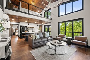 A spacious living area with a high ceiling, a sofa, a coffee table, and large windows offering a view of greenery.