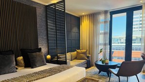 A modern hotel room with a large bed, a sofa, a small table, and a chair. There is a view of a cityscape through the window. - Cape Town Studio 16 on Bree (Cape Town)