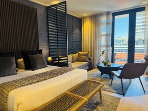 A modern hotel room with a large bed, a sofa, a small table, and a chair. There is a view of a cityscape through the window. - Cape Town Studio 16 on Bree (Cape Town)
