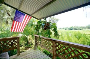 Eine Holzterrasse mit Blick auf ein üppiges Grünfeld, eine amerikanische Flagge und ein überdachter Bereich mit Wellblechdach.