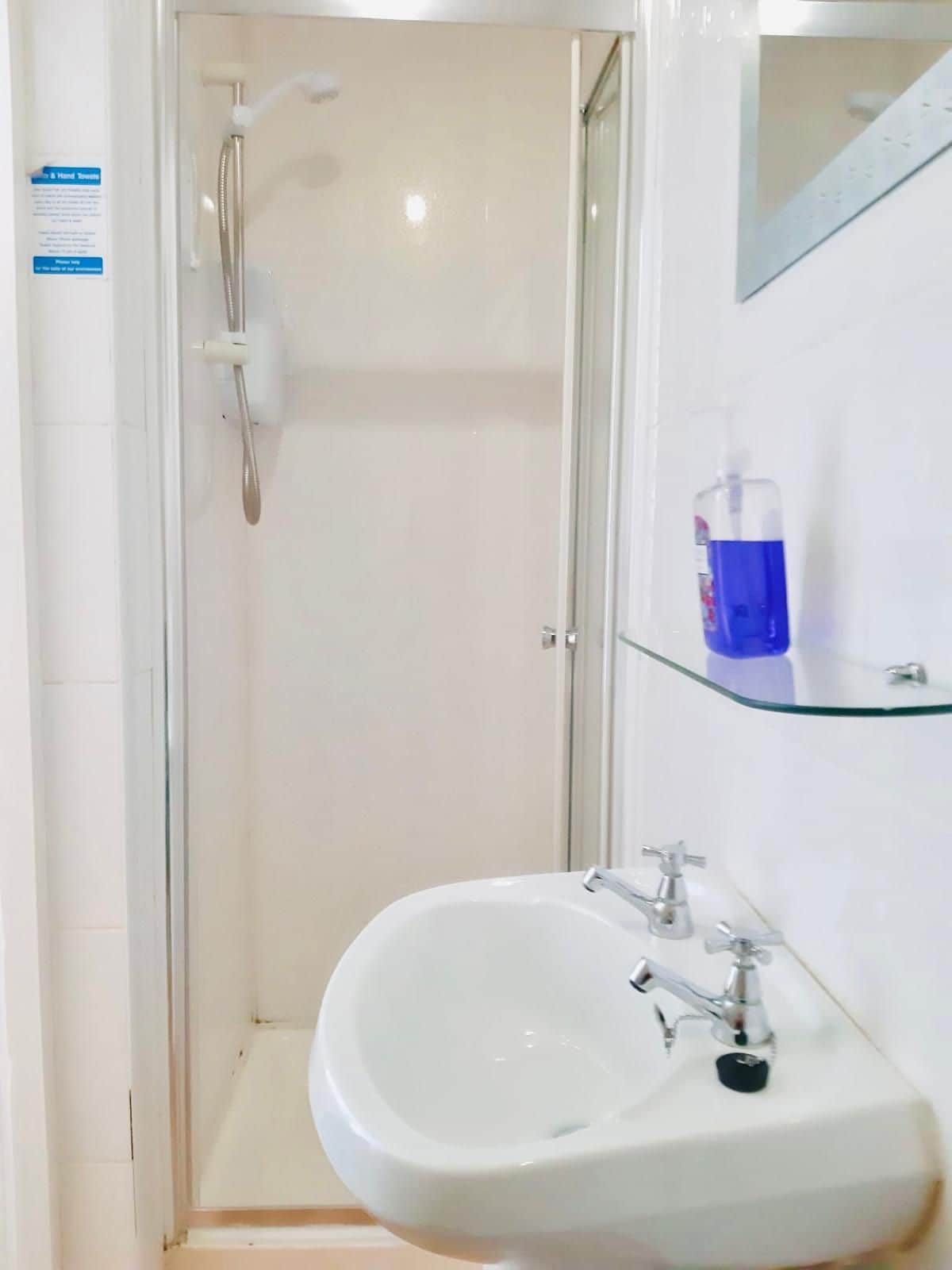 A bathroom with a glass-enclosed shower, a white sink, and a glass shelf with a bottle.