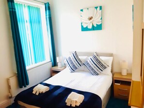 A hotel room with a bed, a nightstand, a lamp, a window with curtains, and a wall-mounted flower painting. - The Beeches Hotel (Blackpool)