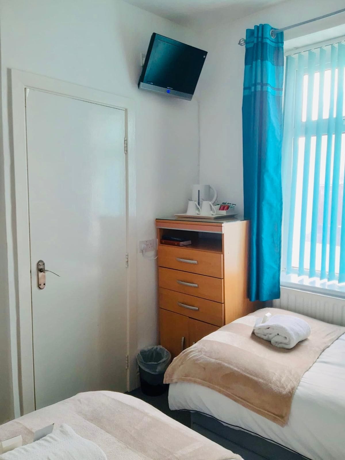 Une chambre à coucher comprenant un lit, une table de chevet avec une bouilloire, un téléviseur fixé au mur, une armoire et une fenêtre avec des rideaux.
