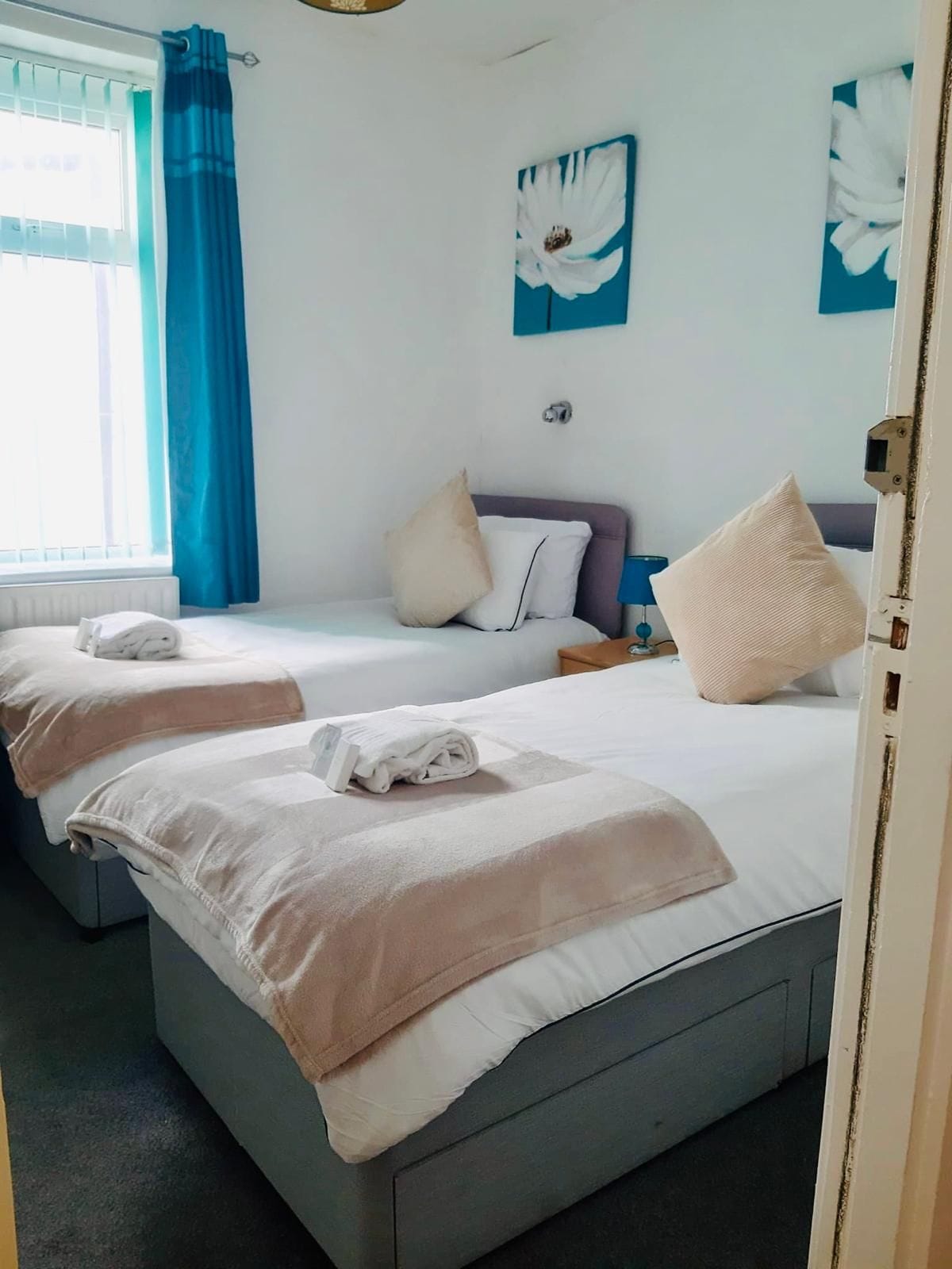 Une chambre d’hôtel avec deux lits, chacun muni d’un oreiller et d’une serviette enroulée. Il y a une fenêtre avec des rideaux bleus, une lampe fixée au mur et un tableau.