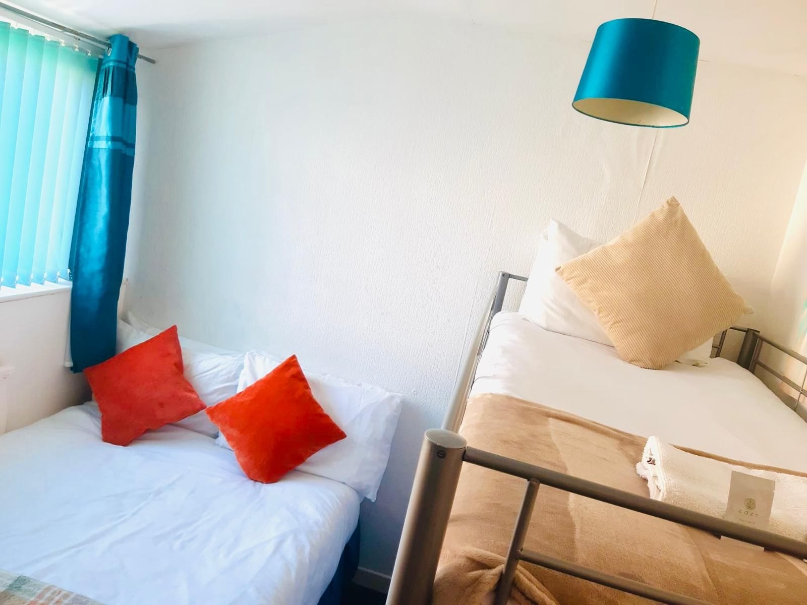 Deux lits avec du linge de lit blanc et des oreillers rouges et beiges, une lampe suspendue bleue et une fenêtre avec des rideaux bleus.