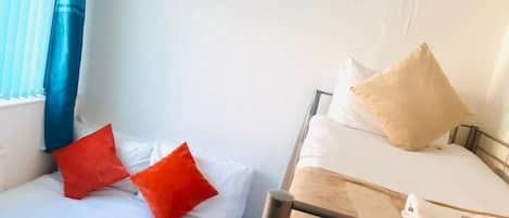 Deux lits avec du linge de lit blanc et des oreillers rouges et beiges, une lampe suspendue bleue et une fenêtre avec des rideaux bleus.