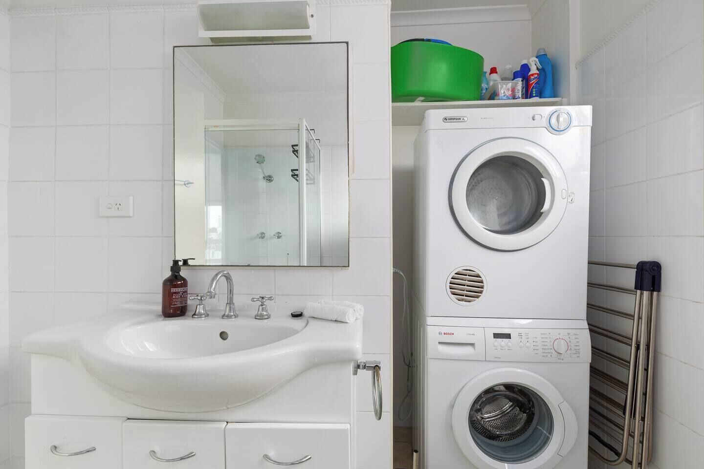 Een badkamer met een wastafel, spiegel, een Bosch wasmachine en een groene container op een plank.