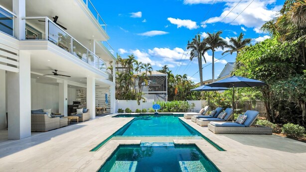 A modern outdoor pool area with a clear blue pool, lounge chairs, and a two-story building with balconies. - Cabana Blu- Luxury Captiva Home with Pool and Steps to the Beach! (Captiva)
