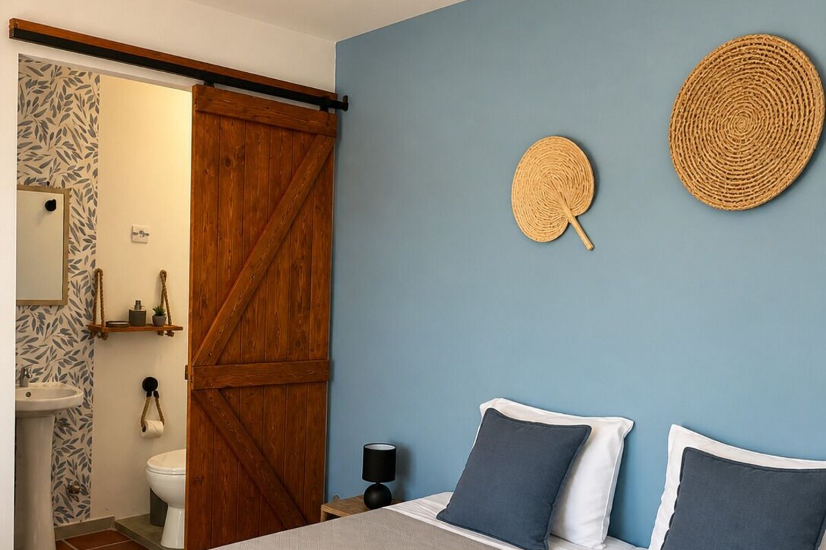 A bedroom with a bed, pillows, a wall-mounted woven item, a wooden door, and a bathroom with a sink and toilet.