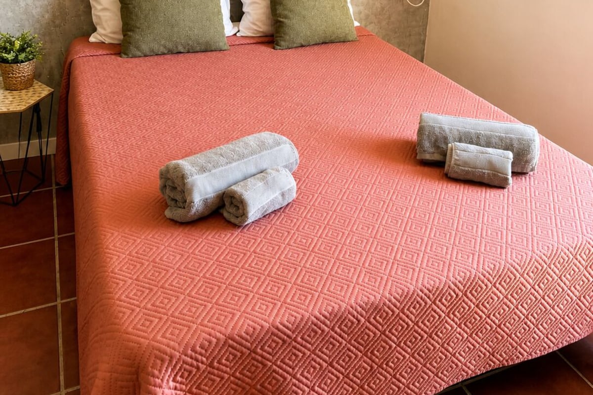 Una cama con una colcha estampada, dos almohadas, un adorno de pared tejido y una toalla enrollada sobre la cama.