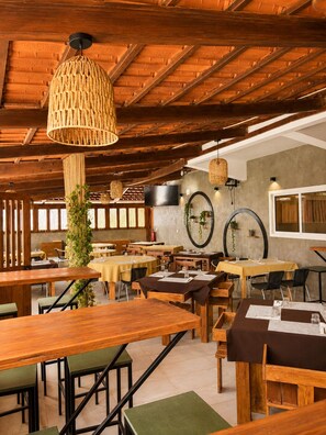 Un restaurante con mesas y sillas de madera, lámparas colgantes tejidas y una pared de piedra con un televisor montado.