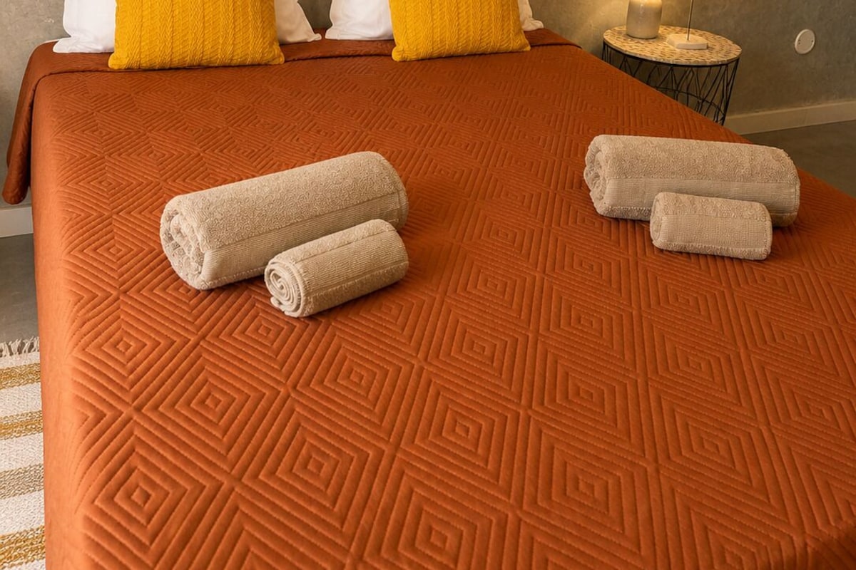 A hotel room with a patterned orange bedspread, two pillows, a small round table with a lamp, and rolled towels on the bed.