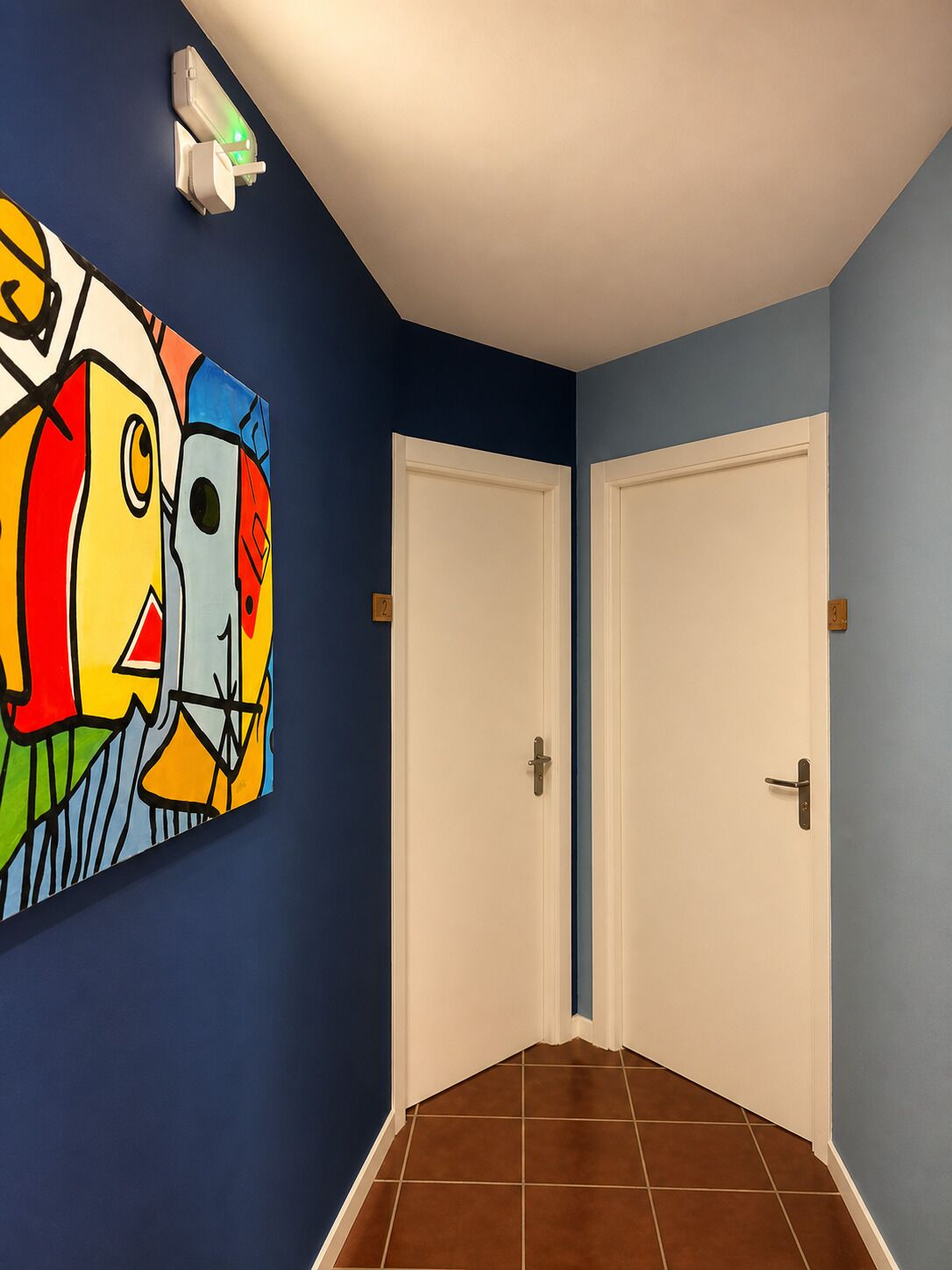 Un couloir avec deux portes blanches, un tableau abstrait aux couleurs vives accroché au mur, et un interrupteur situé au-dessus des portes.