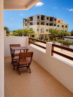 Varanda com mesa e cadeiras de madeira, com vista para um canteiro de obras e um edifício com várias varandas.