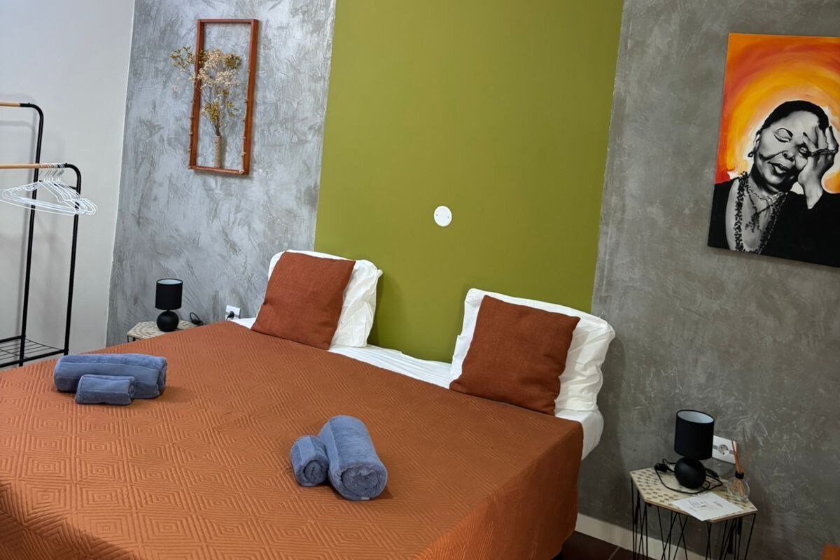 Une chambre à coucher avec un lit, des oreillers, une table de chevet, une lampe, un tableau et une étagère fixée au mur.