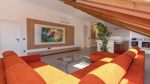 Ein modernes Wohnzimmer mit einem Glastisch, orangefarbenen Sofas, einem großen Fernseher und einer Küche im Hintergrund.
