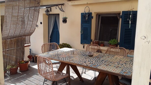 Eine Terrasse im Freien mit einem gefliesten Tisch, Korbstühlen, Blumentöpfen und blauen Fensterläden.
