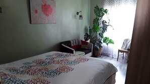 Ein Schlafzimmer mit Bett, Sessel, Sofa, einer Topfpflanze und einer Wandleuchte.