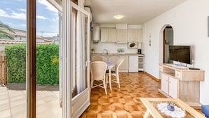 A kitchen with white cabinets, a dining area with wicker chairs, a TV, and a view of a green hedge outside. - 'Bahia Studio' Apartment with Lake Views, Wi-Fi, and Air Conditioning (Empuriabrava)