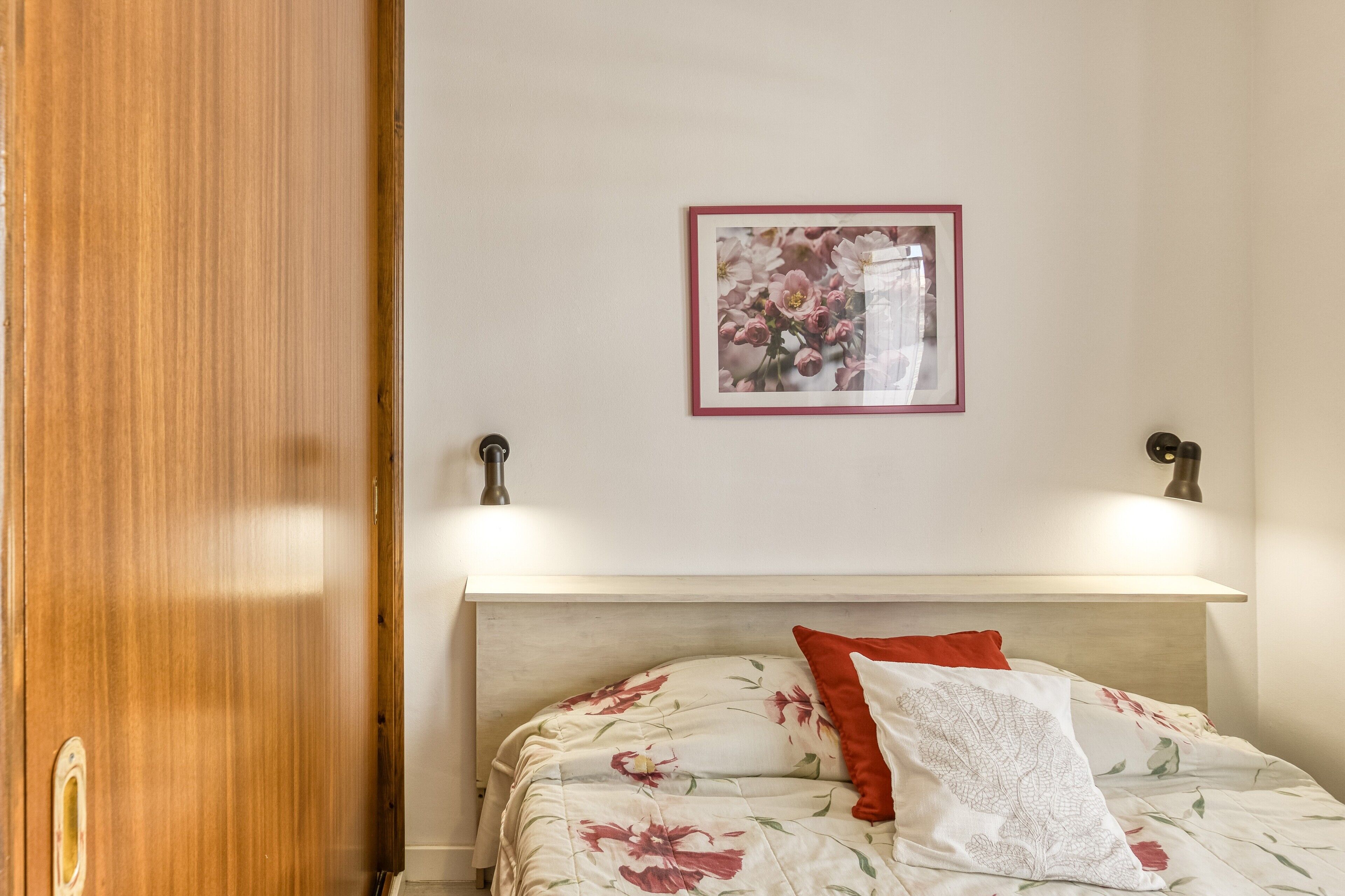 A bedroom with a wooden door, a bed with floral bedding, a framed picture on the wall, and two wall-mounted lamps.