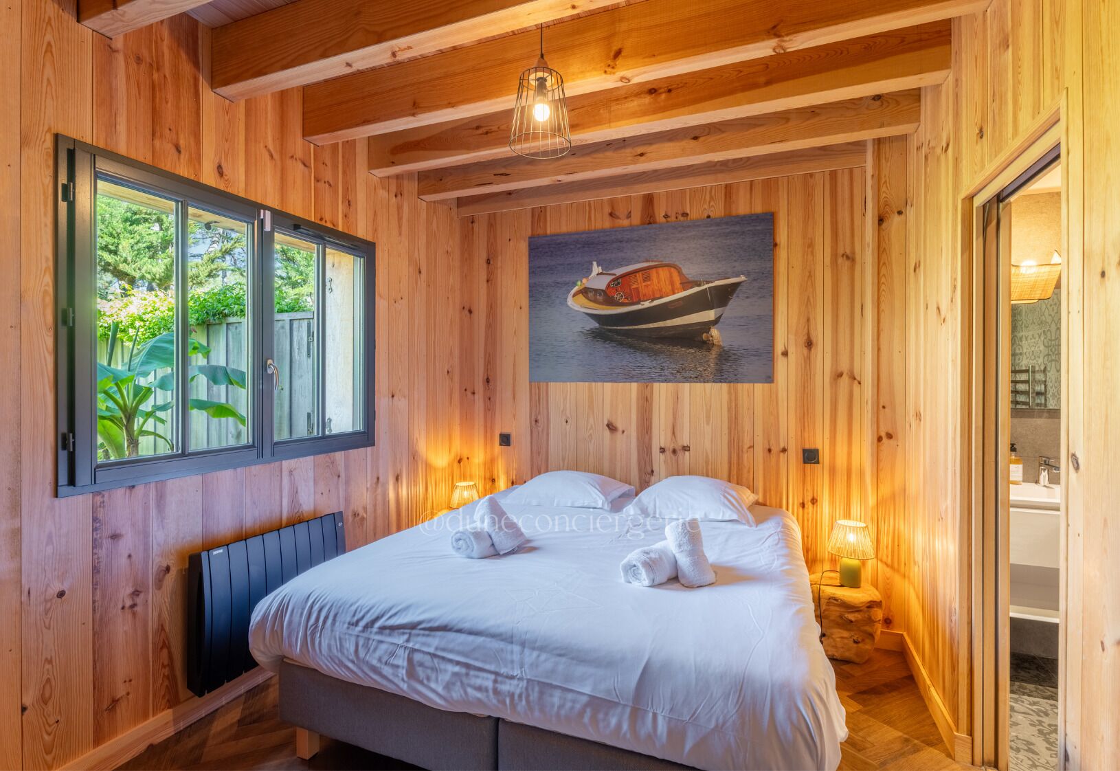 A wooden cabin room with a bed, a window, a painting of a boat, and a bathroom visible through an open door.