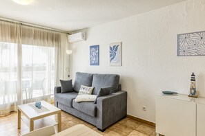 Ein Wohnzimmer mit grauem Sofa, einem kleinen Tisch und einem weißen Schrank. Zwei Wandbilder und eine Klimaanlage sind vorhanden.