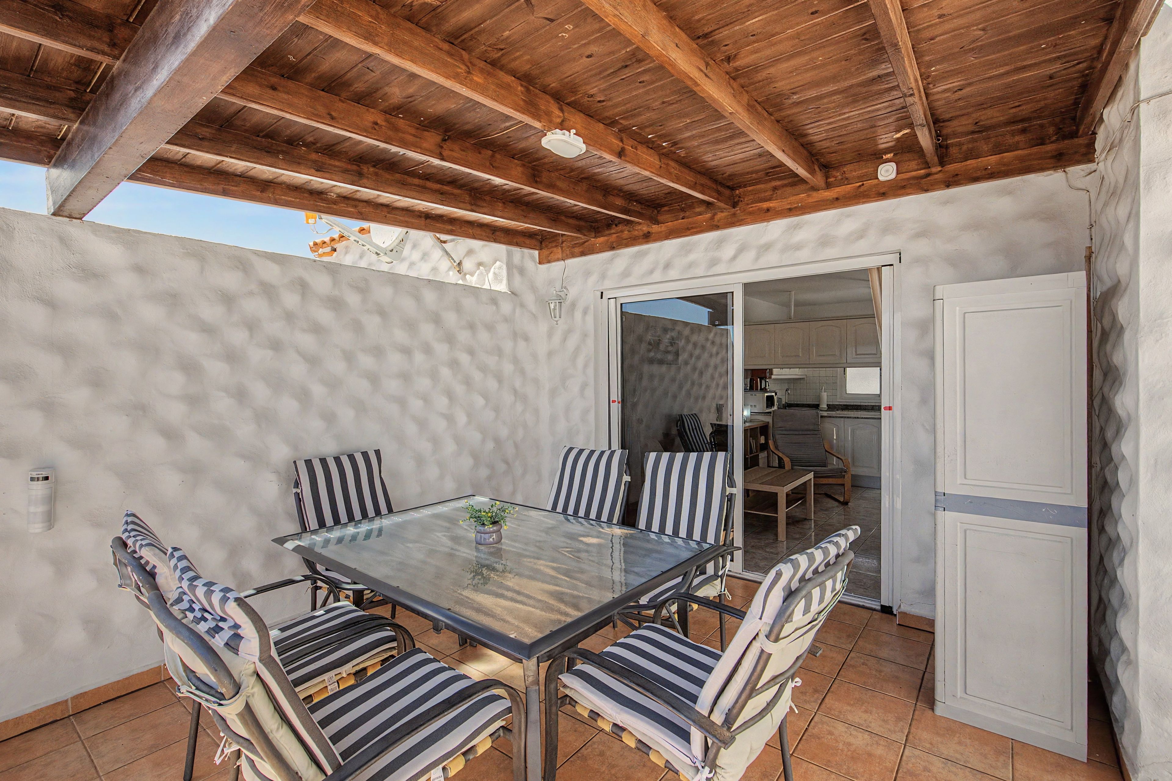An outdoor patio with a glass table surrounded by striped chairs, a wooden ceiling, and a view into a kitchen area.