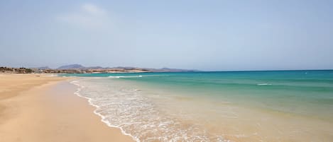 Una playa con agua cristalina, orilla de arena y montañas a lo lejos.