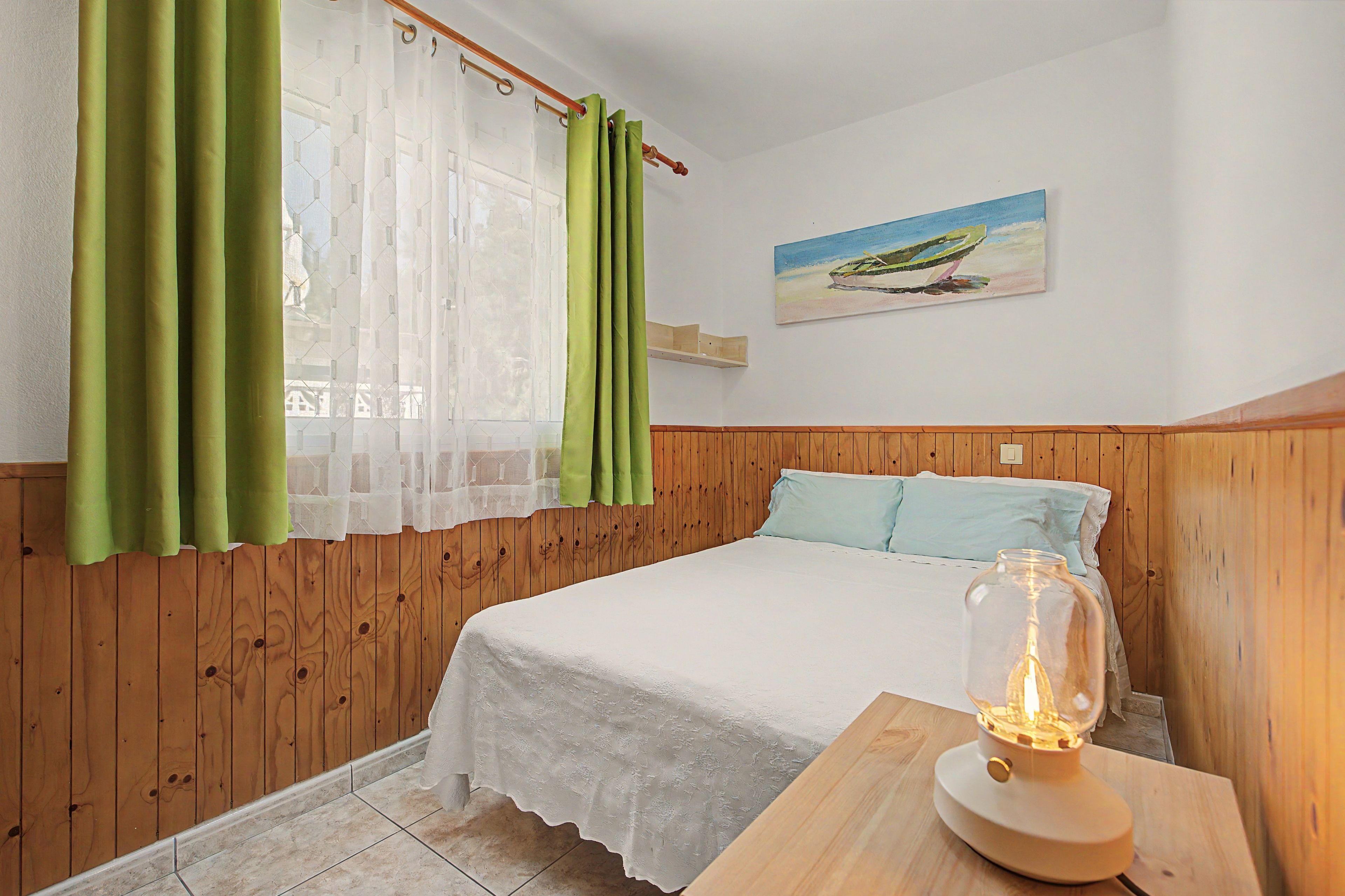 A bedroom with a wooden headboard, a bed with white bedding, green curtains, a painting of a boat on the wall, and a table with a glass jar.