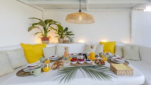 A breakfast setup on a white sofa with a woven table runner, surrounded by potted plants and yellow pillows.