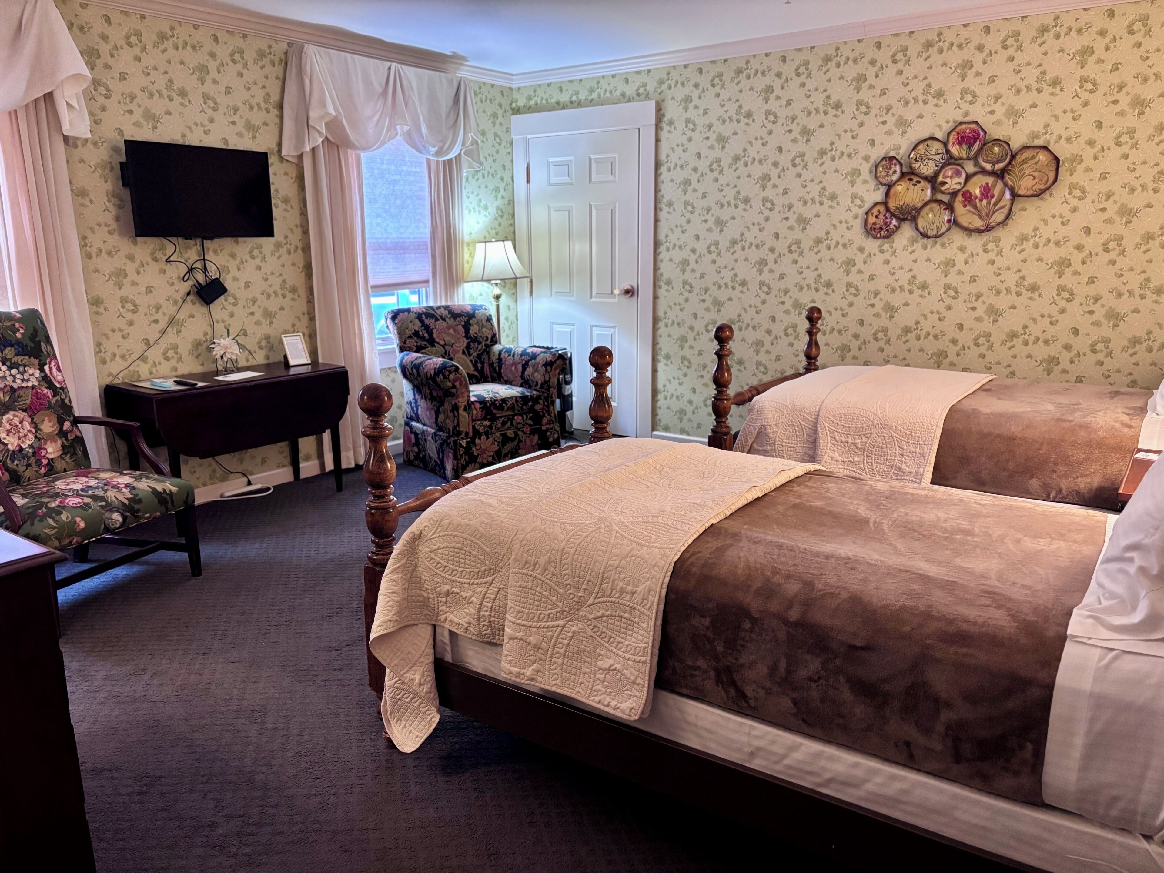 A hotel room with two beds, a desk, a chair, a TV, and a door.