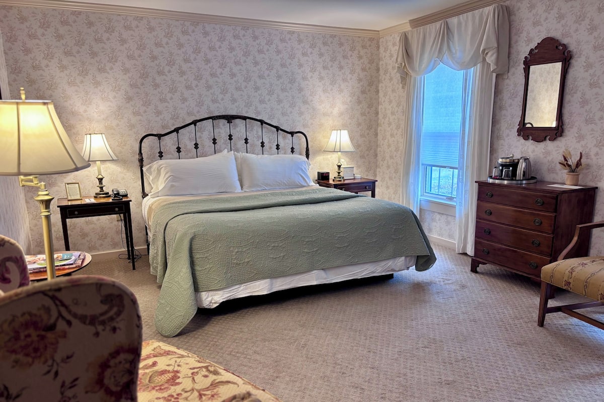 A neatly arranged bedroom with a bed, two bedside tables, a dresser, a chair, and a window with curtains.