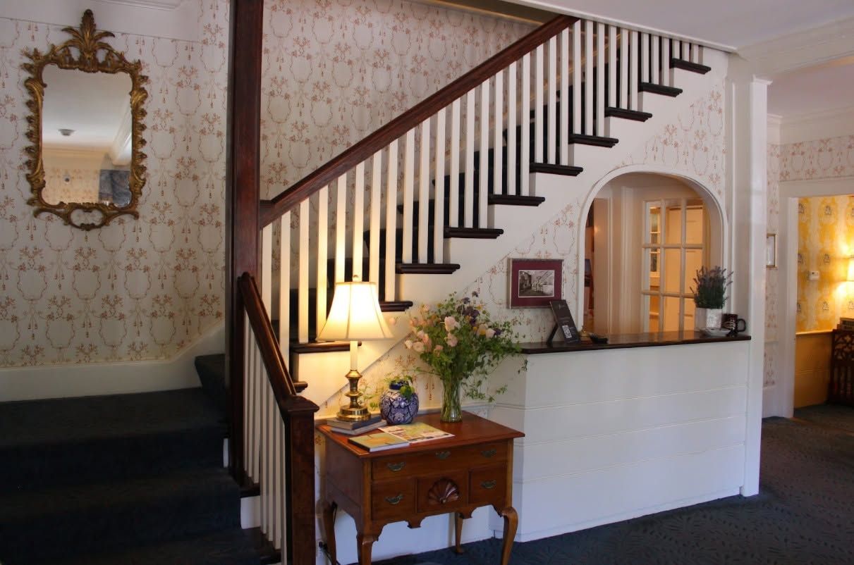 A staircase with a wooden handrail and white balusters, leading to a landing with a desk, a vase with flowers, and a framed picture.
