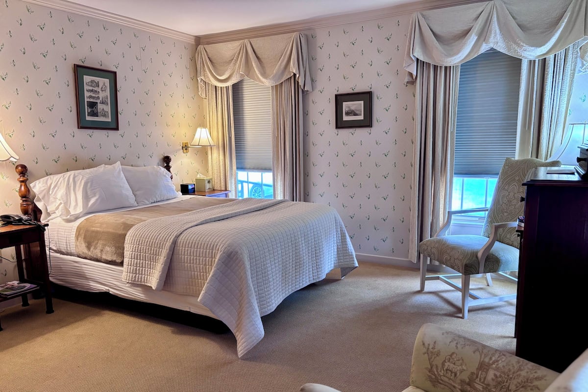 A neatly arranged hotel room with a bed, a chair, a desk, and two framed pictures on the wall.