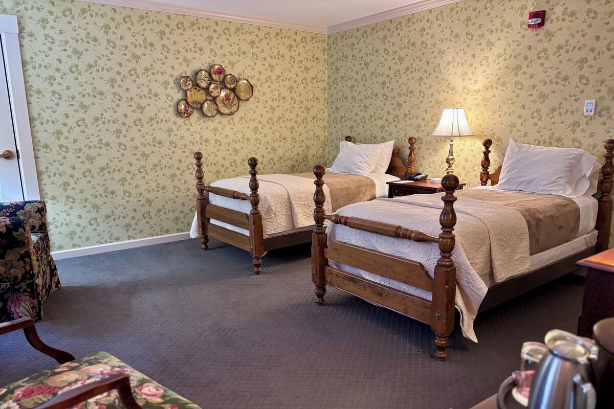 A room with two wooden beds, a floral armchair, a bedside table with a lamp, and a wall clock.