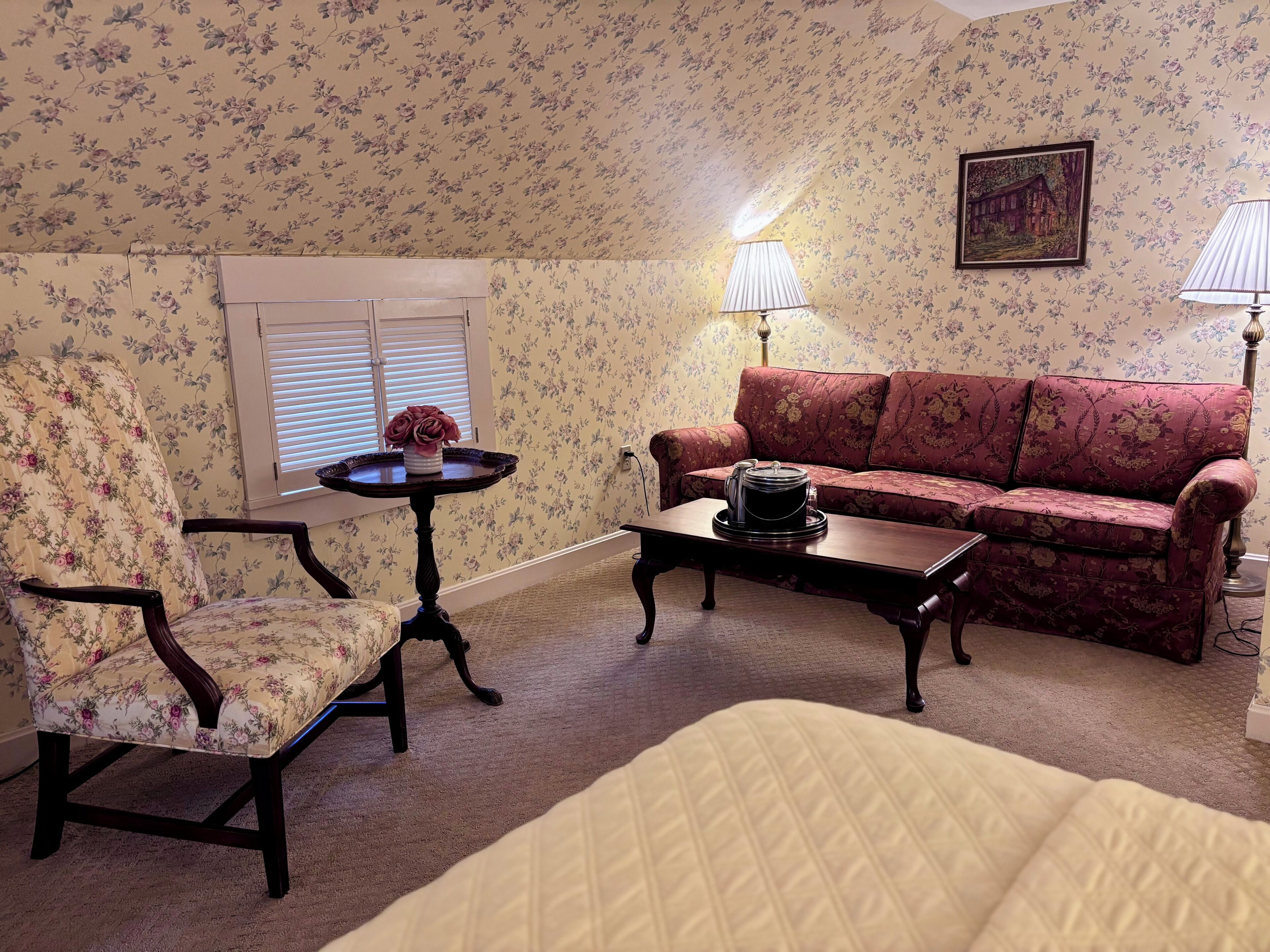 A room with floral wallpaper, a sofa, a chair, a small table with a lamp, and a painting on the wall.
