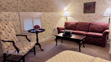 A room with floral wallpaper, a sofa, a chair, a small table with a lamp, and a painting on the wall.