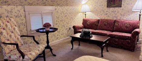 A room with floral wallpaper, a sofa, a chair, a small table with a lamp, and a painting on the wall.