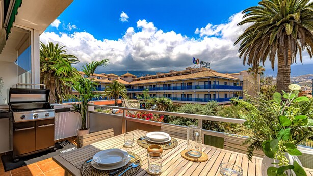 Ein Balkon mit Grill, Esstisch gedeckt mit Tellern und Gläsern sowie Blick auf ein Hotelgebäude mit Palmen.