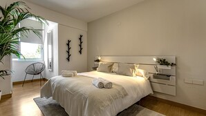 Ein Schlafzimmer mit einem großen Bett, einem Nachttisch, einem Sessel, einer Pflanze und einem Fenster.