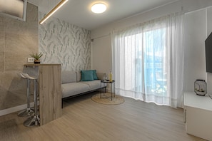 Ein modernes Wohnzimmer mit einer Couch, einem kleinen runden Tisch, einem Fernseher und einem Fenster mit durchscheinenden Vorhängen.