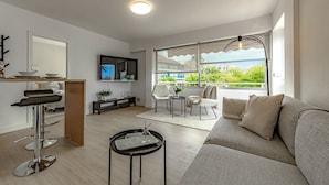 Un salon moderne comprenant un canapé, un coin repas, un téléviseur et une grande fenêtre donnant sur la ville.