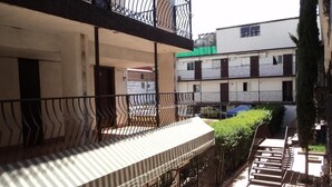 Un immeuble résidentiel à plusieurs étages, doté de balcons, d’une galerie couverte et d’une haie verdoyante.