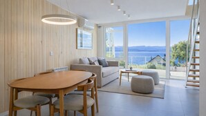 A modern living room with a wooden dining table, sofa, and ottoman, large windows with a view of the sea, and a staircase leading to an upper level. - Signature Lake View, Luxury Residences - Unit B06 (San Carlos de Bariloche)