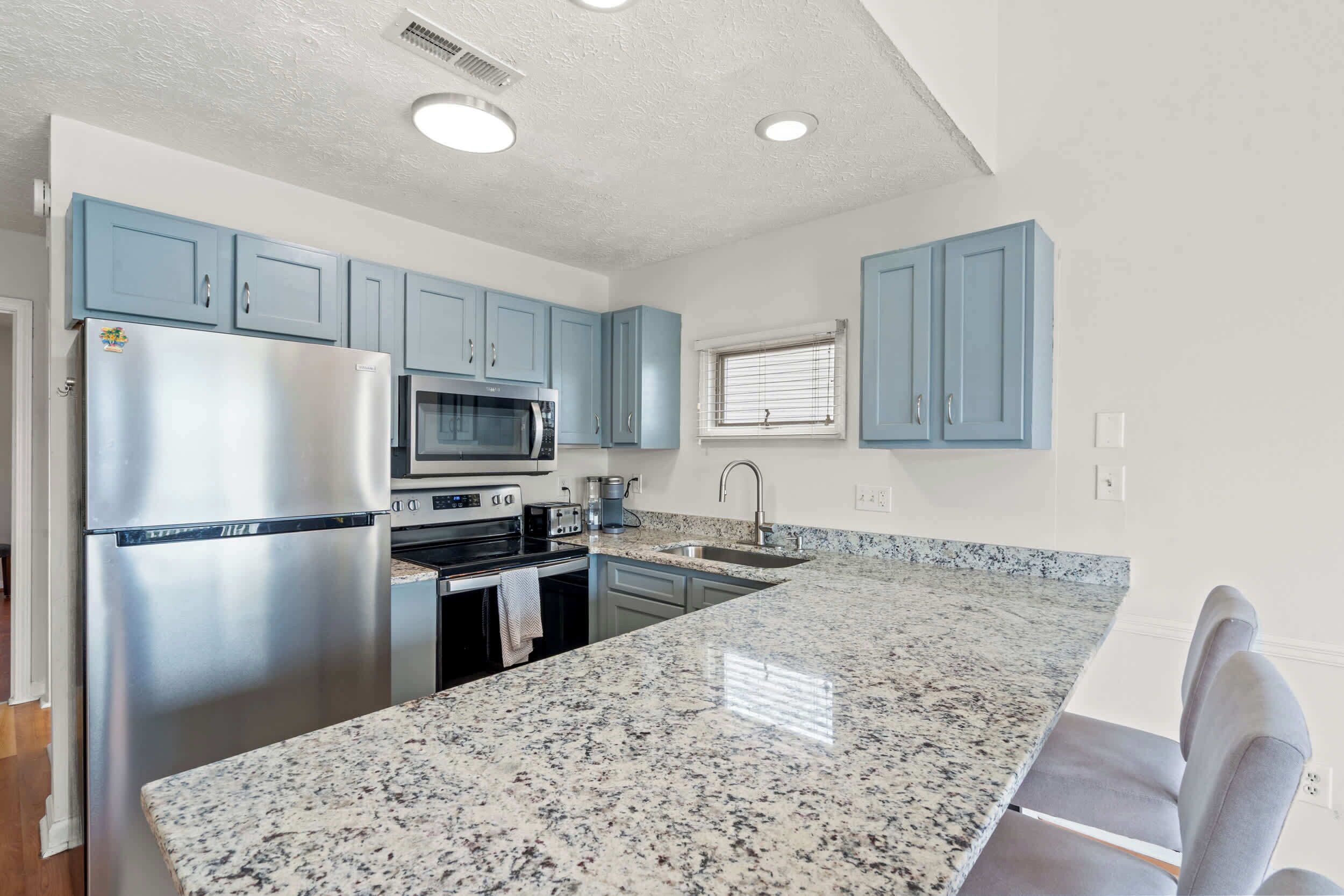 A kitchen with blue cabinets, stainless steel appliances, granite countertops, and a window with blinds.