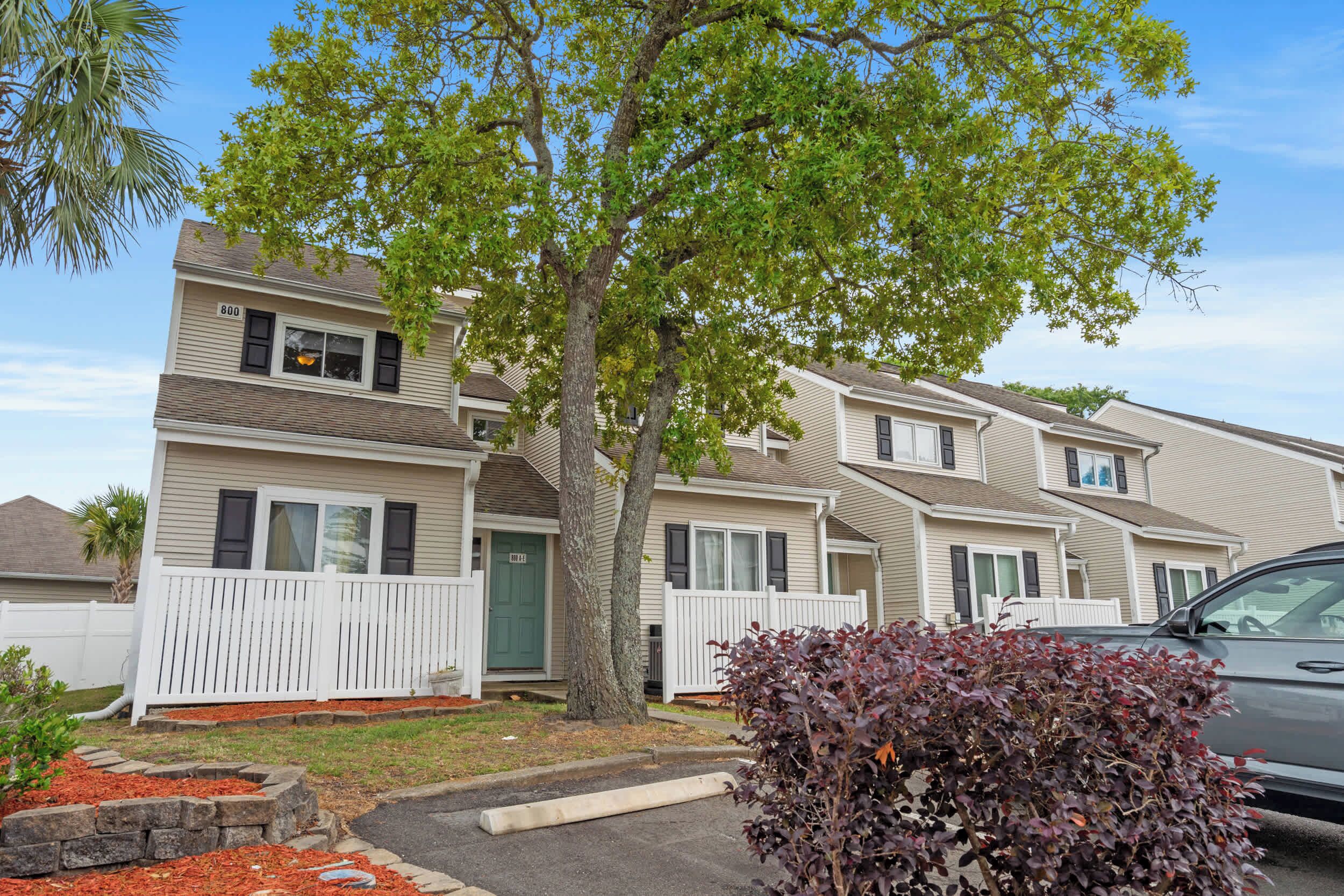 A row of two-story townhouses with a white picket fence, a tree in front, and a parked car.