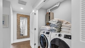 A laundry room with two front-loading washing machines, a mirror with a woven frame, and a stack of folded towels. - Luxury 3BR with Seaside Views Jersey Shore Gem (Seaside Heights)