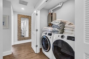 A laundry room with two front-loading washing machines, a mirror with a woven frame, and a stack of folded towels. - Luxury 3BR with Seaside Views Jersey Shore Gem (Seaside Heights)