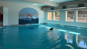 Ein Hallenbad mit Wandgemälde einer Berglandschaft, großen Fenstern und einer schwimmenden Person.