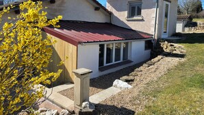 Una casa con tejado rojo, chimenea de piedra y un pequeño jardín con grava y un árbol floreciente de color amarillo.